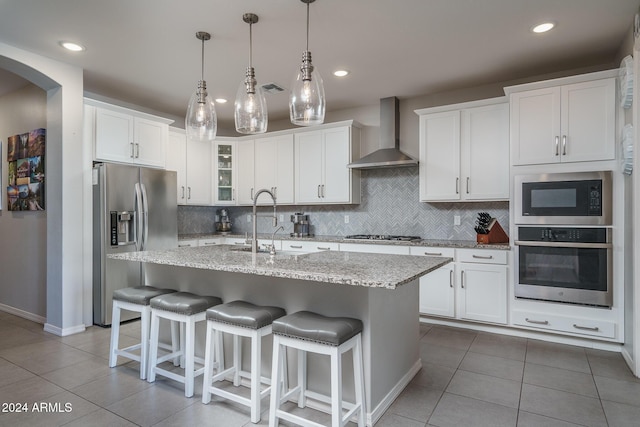 kitchen with an island with sink, white cabinets, stainless steel appliances, and wall chimney range hood