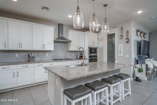 kitchen with white cabinetry, hanging light fixtures, wall chimney range hood, an island with sink, and appliances with stainless steel finishes