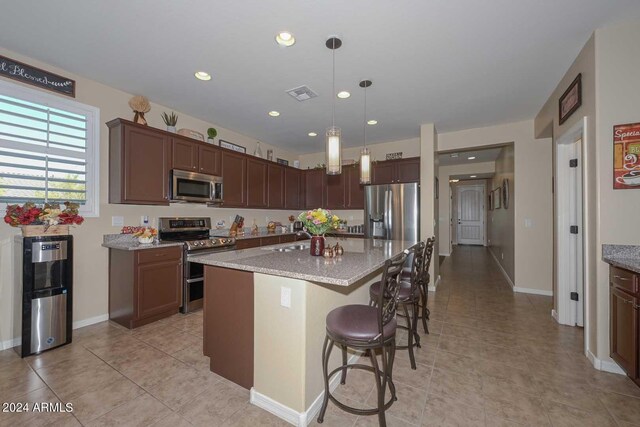 kitchen featuring appliances with stainless steel finishes, hanging light fixtures, light stone counters, a center island, and dark brown cabinetry