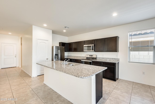 kitchen featuring light tile patterned floors, light stone counters, stainless steel appliances, a sink, and an island with sink