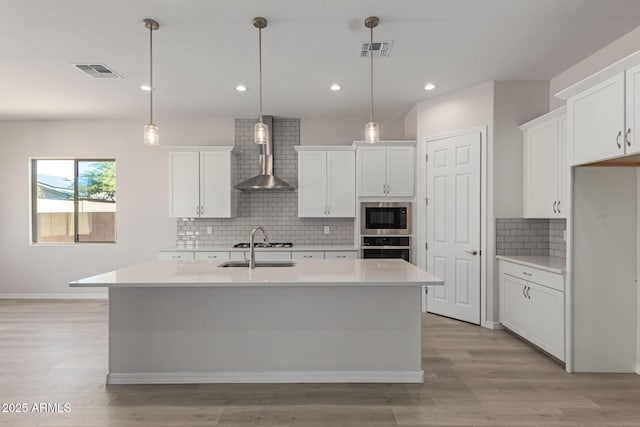 kitchen with a center island with sink, built in microwave, white cabinets, wall chimney range hood, and decorative light fixtures