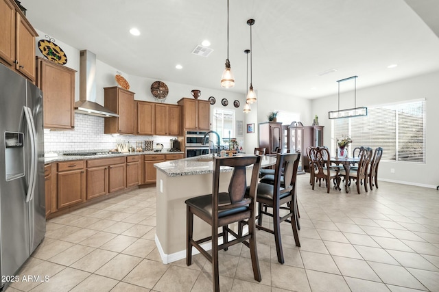 kitchen featuring wall chimney exhaust hood, appliances with stainless steel finishes, a kitchen breakfast bar, an island with sink, and light stone countertops