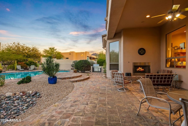 patio terrace at dusk featuring a fenced in pool, grilling area, ceiling fan, and exterior fireplace