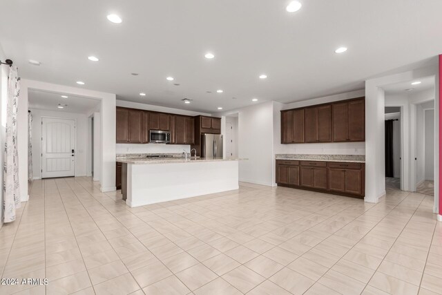 kitchen with light stone counters, stainless steel appliances, a center island with sink, and dark brown cabinetry