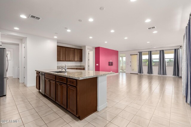 kitchen with light tile patterned floors, stainless steel appliances, light stone counters, sink, and a center island with sink