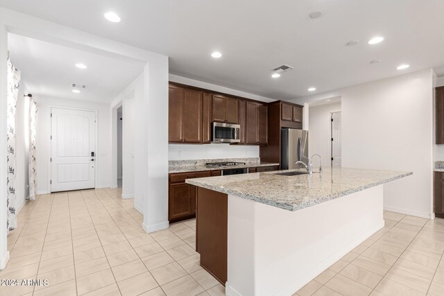 kitchen with appliances with stainless steel finishes, light stone countertops, sink, dark brown cabinets, and a center island with sink