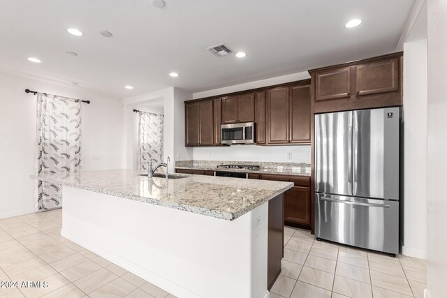 kitchen with dark brown cabinets, appliances with stainless steel finishes, an island with sink, light stone counters, and light tile patterned flooring