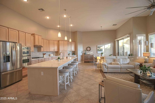 kitchen featuring appliances with stainless steel finishes, hanging light fixtures, an island with sink, a kitchen bar, and light brown cabinetry