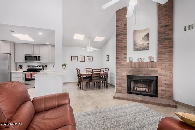 living room with a skylight, high vaulted ceiling, a brick fireplace, light tile patterned floors, and ceiling fan