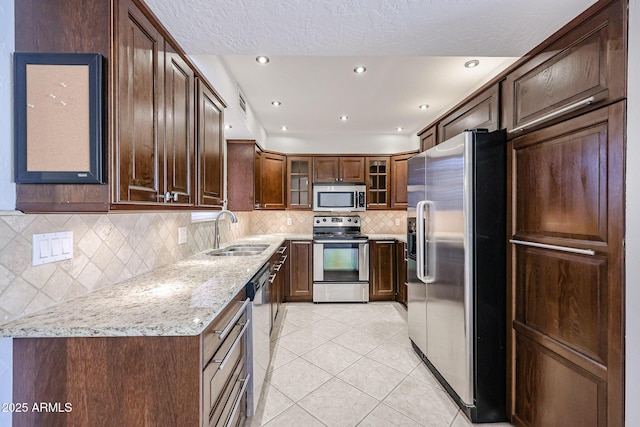 kitchen featuring a sink, light tile patterned floors, appliances with stainless steel finishes, light stone countertops, and glass insert cabinets