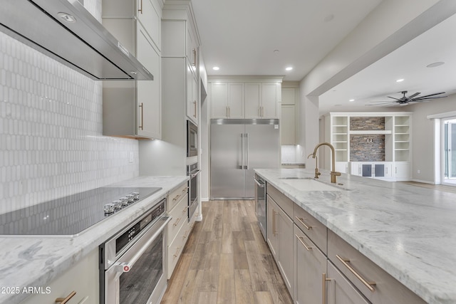 kitchen featuring light wood-style flooring, light stone counters, built in appliances, wall chimney range hood, and a sink