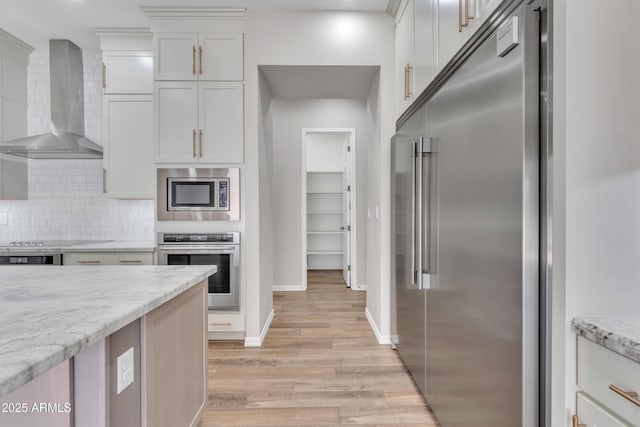 kitchen featuring light stone counters, decorative backsplash, built in appliances, light wood-type flooring, and wall chimney exhaust hood