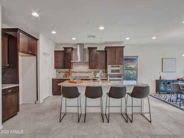 kitchen with sink, stainless steel appliances, light stone countertops, a center island with sink, and wall chimney exhaust hood