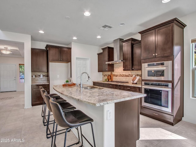 kitchen featuring sink, a kitchen island with sink, light stone counters, tasteful backsplash, and wall chimney exhaust hood