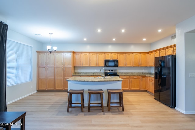 kitchen featuring black appliances, a kitchen island with sink, light stone counters, hanging light fixtures, and light hardwood / wood-style flooring