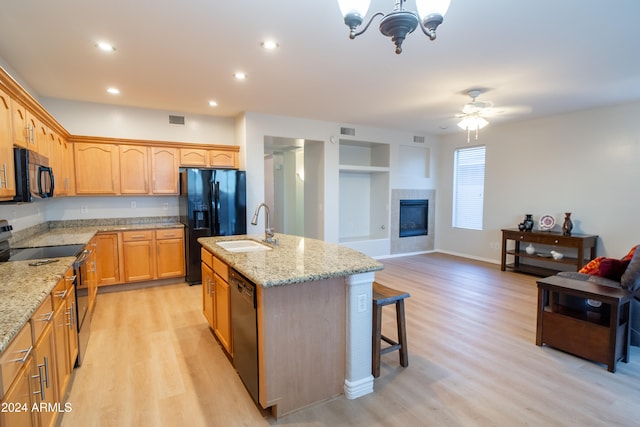 kitchen featuring a center island with sink, light stone counters, black appliances, sink, and light wood-type flooring