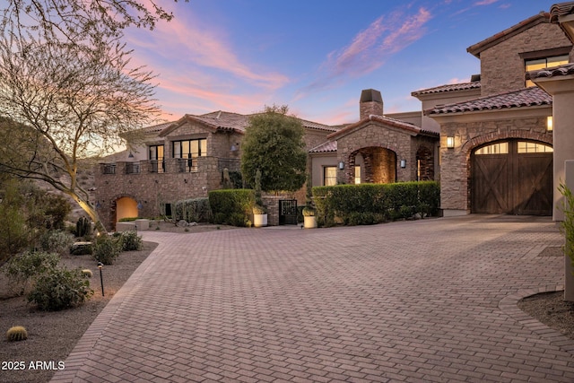 mediterranean / spanish house featuring stone siding, a tile roof, a chimney, decorative driveway, and stucco siding