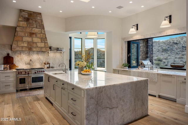 kitchen featuring sink, light stone counters, double oven range, pendant lighting, and a kitchen island with sink