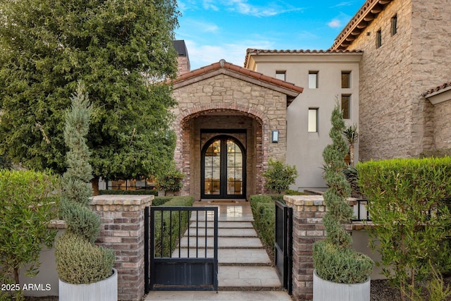 entrance to property with stone siding, french doors, a tiled roof, a gate, and stucco siding