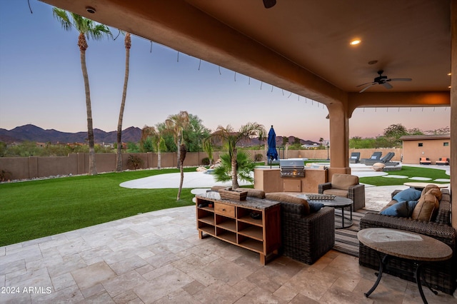 patio terrace at dusk featuring ceiling fan, a mountain view, a grill, a yard, and an outdoor kitchen