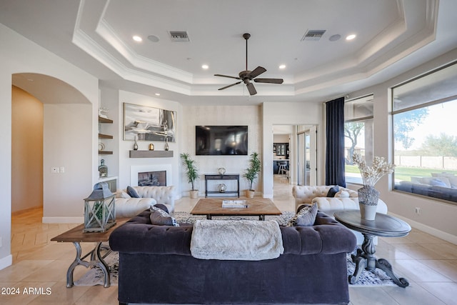 tiled living room featuring a raised ceiling, a fireplace, ceiling fan, built in features, and ornamental molding