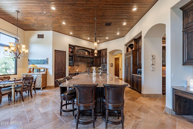 kitchen with light stone counters, a kitchen island with sink, hanging light fixtures, and wooden ceiling