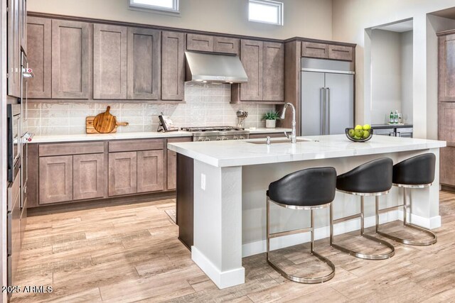 kitchen with a sink, backsplash, built in fridge, light wood-style floors, and wall chimney range hood