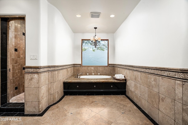 bathroom with an inviting chandelier, wainscoting, tile walls, a garden tub, and stone tile flooring