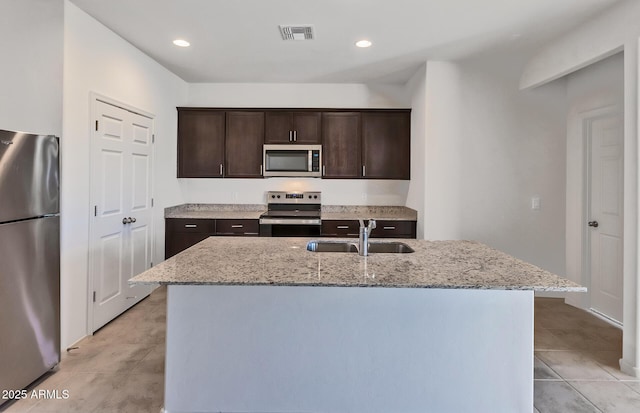 kitchen with appliances with stainless steel finishes, an island with sink, sink, light stone countertops, and dark brown cabinets