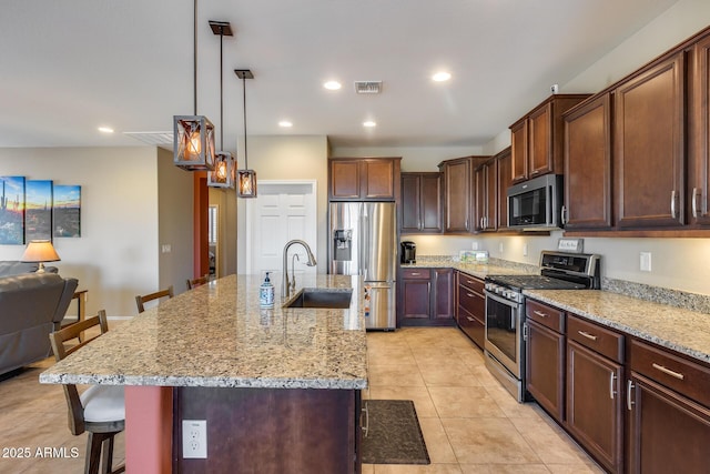 kitchen with hanging light fixtures, stainless steel appliances, light stone countertops, an island with sink, and a kitchen bar