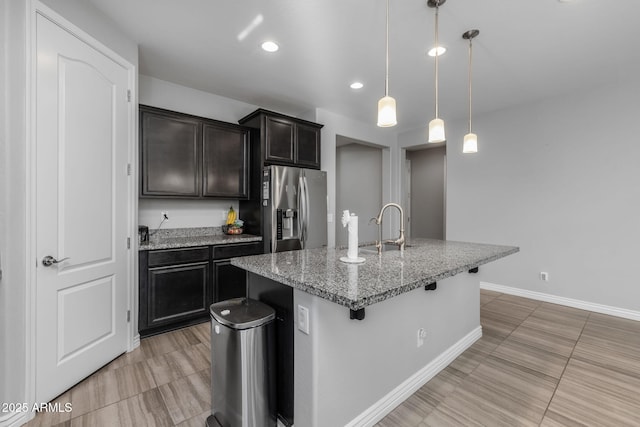 kitchen with sink, light stone counters, stainless steel refrigerator with ice dispenser, decorative light fixtures, and a kitchen island with sink