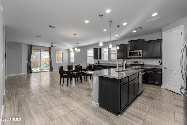 kitchen featuring light stone counters, ceiling fan with notable chandelier, stainless steel appliances, a kitchen island with sink, and hanging light fixtures