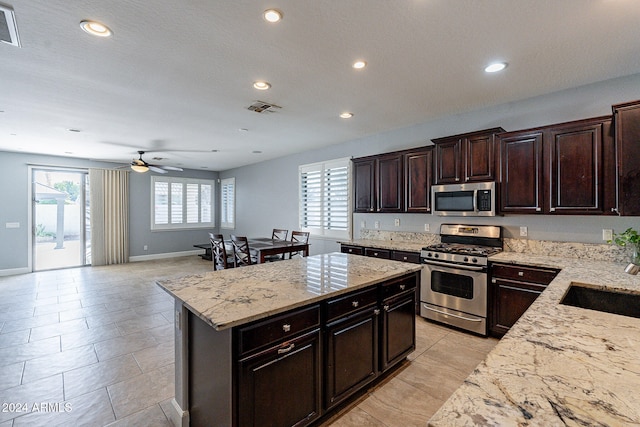 kitchen featuring ceiling fan, dark brown cabinets, appliances with stainless steel finishes, a center island, and light stone countertops