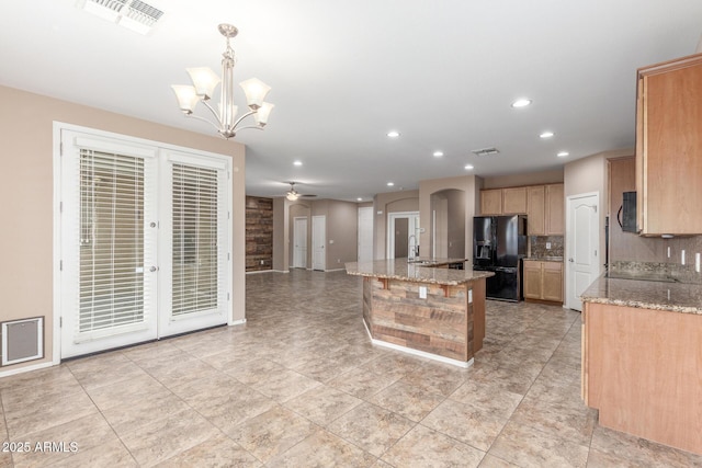 kitchen featuring black fridge, light stone counters, hanging light fixtures, a center island with sink, and decorative backsplash