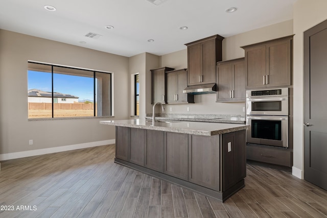 kitchen featuring stainless steel double oven, dark hardwood / wood-style flooring, sink, black electric cooktop, and an island with sink