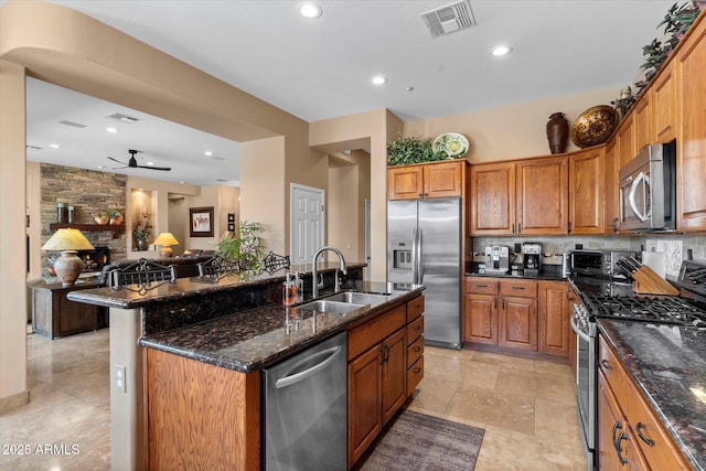 kitchen featuring stainless steel appliances, dark stone countertops, an island with sink, sink, and ceiling fan