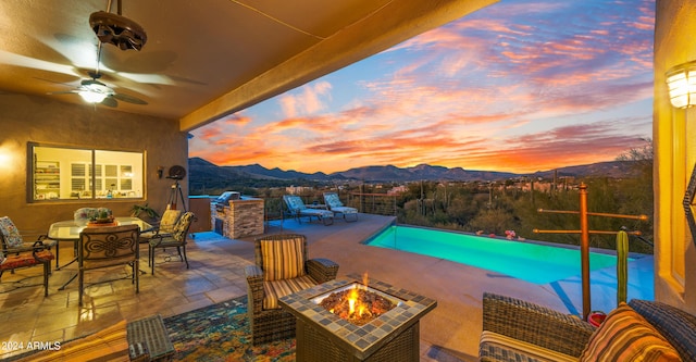 pool at dusk with a mountain view, ceiling fan, a patio area, and an outdoor fire pit