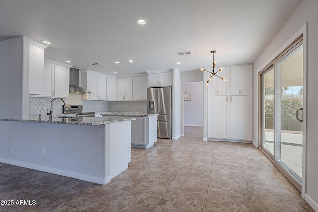 kitchen with wall chimney exhaust hood, stainless steel appliances, stone countertops, an inviting chandelier, and white cabinetry