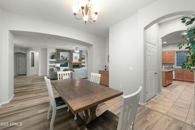 dining space with baseboards, arched walkways, light wood-type flooring, a fireplace, and ceiling fan with notable chandelier