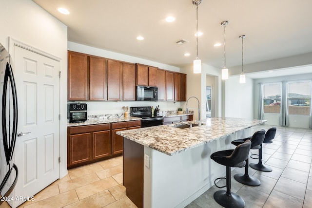 kitchen featuring light stone countertops, a kitchen island with sink, black appliances, sink, and hanging light fixtures