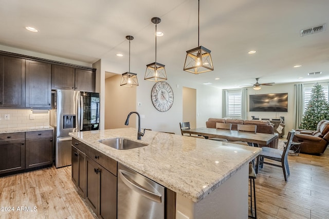 kitchen with light stone counters, stainless steel appliances, sink, decorative light fixtures, and a center island with sink