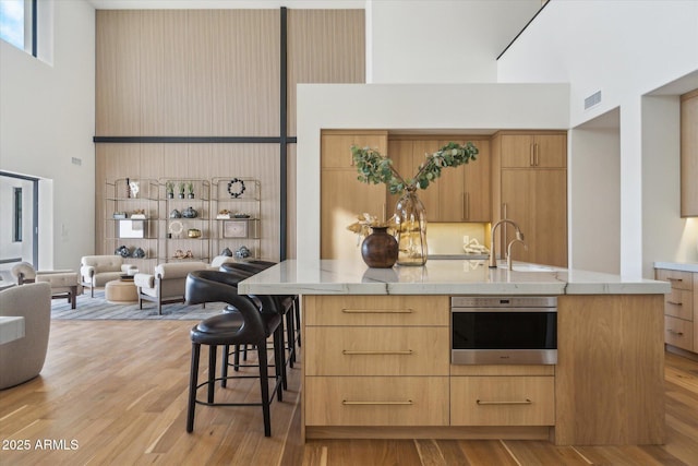 kitchen featuring oven, light hardwood / wood-style flooring, a center island with sink, light brown cabinetry, and a high ceiling