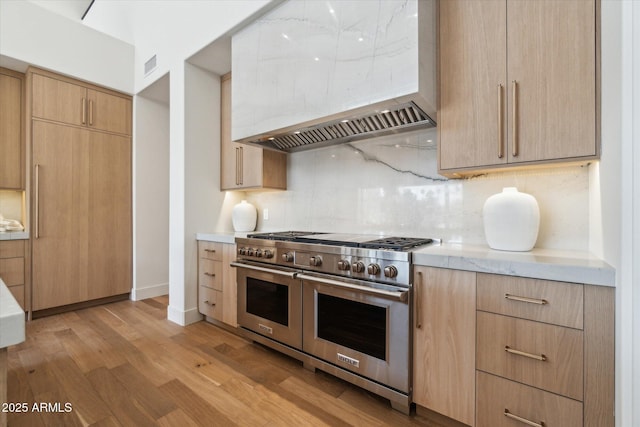 kitchen featuring light wood-type flooring, premium range hood, double oven range, light brown cabinets, and backsplash