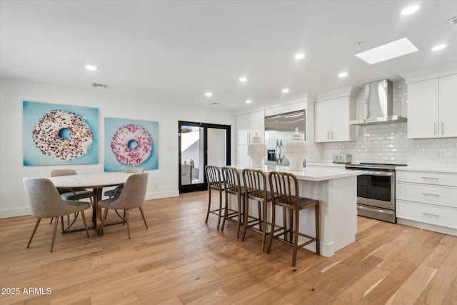 kitchen featuring wall chimney exhaust hood, stainless steel appliances, an island with sink, decorative backsplash, and white cabinetry