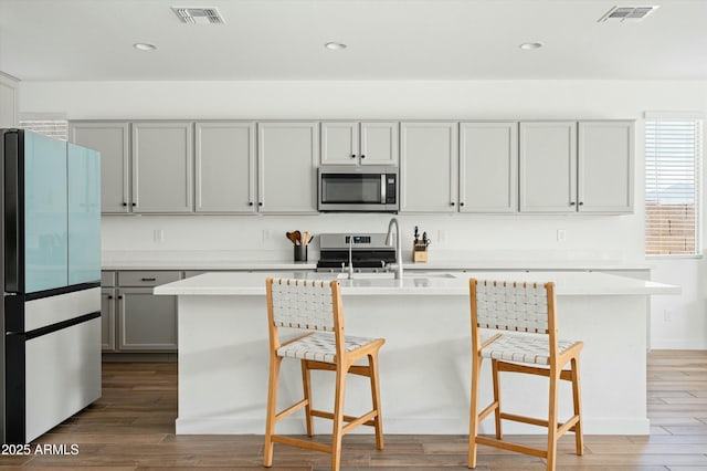 kitchen with wood-type flooring, stainless steel appliances, a center island with sink, and gray cabinetry