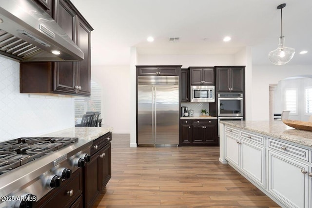 kitchen with pendant lighting, white cabinetry, exhaust hood, built in appliances, and light hardwood / wood-style flooring