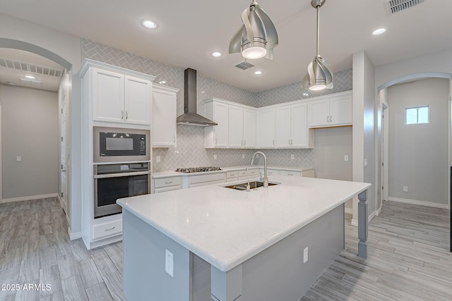 kitchen with sink, wall chimney exhaust hood, an island with sink, white cabinets, and appliances with stainless steel finishes