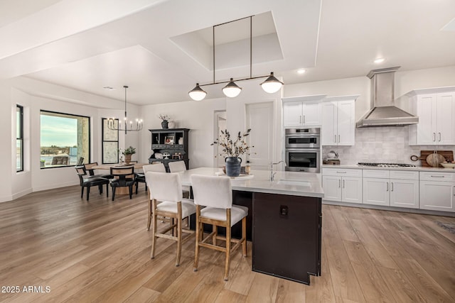 kitchen with hanging light fixtures, an island with sink, wall chimney range hood, and white cabinets