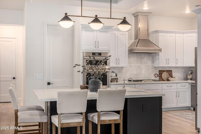 kitchen with white cabinetry, appliances with stainless steel finishes, an island with sink, pendant lighting, and wall chimney range hood