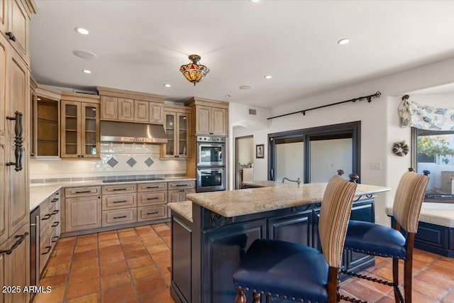 kitchen featuring a breakfast bar, under cabinet range hood, stainless steel double oven, decorative backsplash, and black electric stovetop